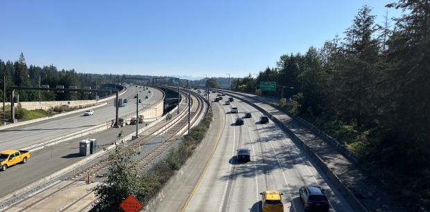 An aerial shot of I-90 on the East Channel Bridge with cars traveling, surrounded by trees in the background under a clear blue sky.