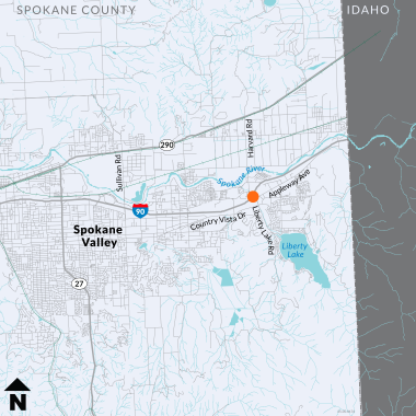 A map showing the region around Spokane Valley and Liberty Lake. The map uses shades of blue and grey to distinguish different geographical and man-made features. The Spokane River flows horizontally across the upper section, highlighted in light blue, while the nearby city of Liberty Lake appears in the lower right area. An orange dot is visible on Liberty Lake Road, indicating location of ramp meters.