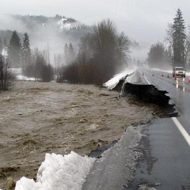A highway washed out by a flooded river next to orange cones.