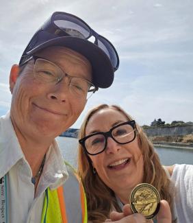 Two people taking a selfie near water, with one holding a gold coin.