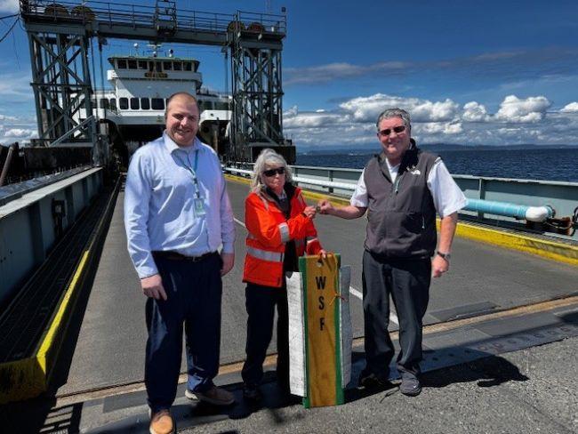 Three people standing on the dock at Fauntleroy terminal with a ferry in the background. and person in middle holding up a piece of wood