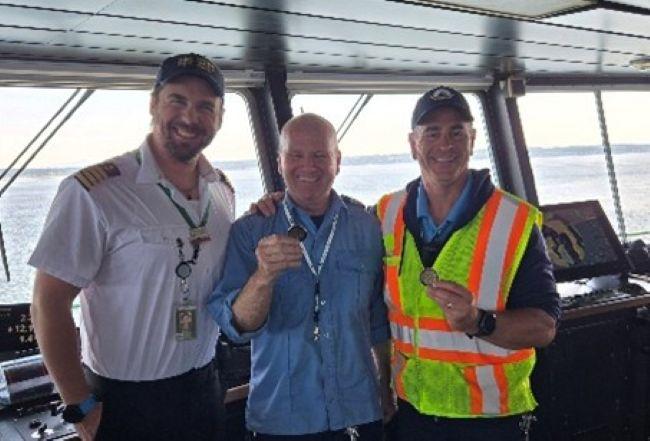 Three people standing in a ferry's pilothouse with two holding up coins.