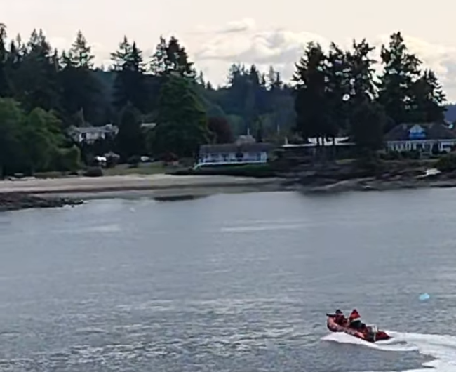 A rescue boat with people on a waterway, surrounded by evergreen trees and homes.