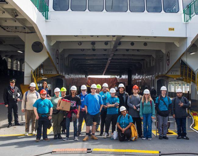 A group wearing safety helmets stands on a ferry deck in front of an open loading area.