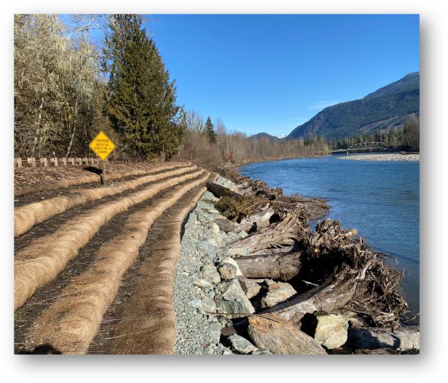 Riverbank with nature-based erosion controls along a highway with a distant bridge and mountains.